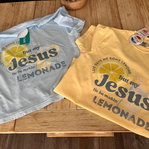 Può includere: Due magliette a maniche corte, una azzurra e una gialla, con la scritta "Life gave me some lemons but my Jesus He be makin' lemonade." Le magliette hanno una grafica di una fetta di limone e limonata.