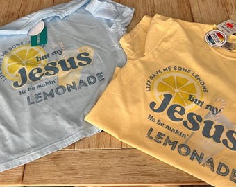 Puede incluir: Dos camisetas de manga corta, una azul claro y otra amarilla, con el texto "Life gave me some lemons but my Jesus He be makin' lemonade." Las camisetas tienen un gráfico de una rodaja de limón y limonada.