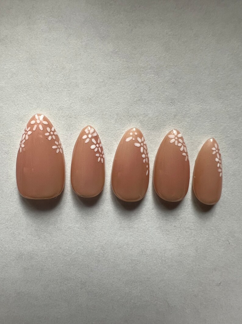 Op de afbeelding: Set van zes nudekleurige kunstnagels met witte bloemenmotieven.