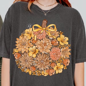 May include: Dark gray t-shirt featuring a floral pumpkin design. The pumpkin is composed of various flowers in shades of orange, yellow, and brown, with a yellow bow and stem. The shirt has a crew neck and short sleeves.