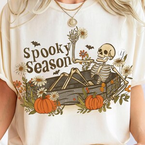 May include: Cream-colored t-shirt featuring a skeleton in a coffin surrounded by pumpkins and flowers. The text "Spooky Season" is printed above the skeleton. Bats are also included in the design.