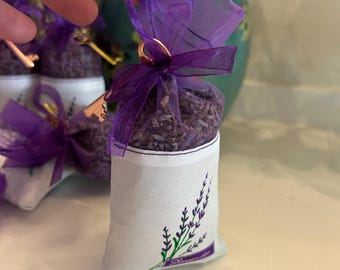 May include: A small white sachet filled with dried lavender, tied with a purple ribbon and a decorative key. The sachet has a lavender sprig design in green and purple. The sachet is held in a hand.