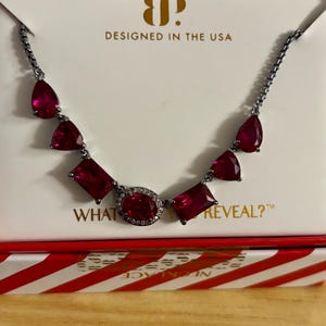 May include: A necklace featuring a dark metal chain and seven red faceted stones in pear, triangle, and rectangular shapes. The central oval stone is surrounded by small clear crystals. The packaging displays the text DESIGNED IN THE USA and WHAT WILL YOU REVEAL?