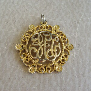 May include: A gold-toned pendant with a delicate filigree design featuring a monogram in the center. The pendant is made of metal and has a loop at the top for attaching a chain.