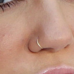 May include: A close-up of a small, thin, gold nose ring. The ring is a simple hoop shape and is worn in the left nostril.
