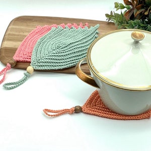 Pode incluir: Dois pegadores de panela de crochê em rosa e verde com contas de madeira nos laços de suspensão. Uma panela de cerâmica branca com uma borda dourada está sobre um terceiro pegador de panela de crochê em pêssego.