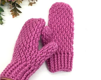 Peut inclure: Une paire de mitaines tricotées à la main, de couleur rose vif. Les mitaines présentent un motif de point texturé et sont conçues pour la chaleur et le confort. L'image montre les mitaines sur fond blanc, avec des accents floraux en haut à gauche.