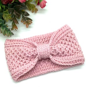 Puede incluir: Una diadema de punto rosa claro con un detalle de lazo. La diadema está hecha de un hilo suave y texturizado y está diseñada para mantener las orejas calientes. El lazo está centrado en la diadema. Flores rosas en el fondo.