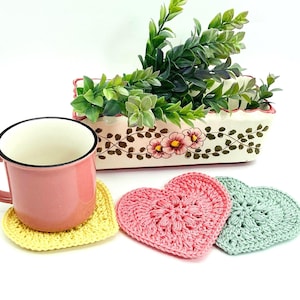 Peut inclure: Trois dessous-de-verre en forme de cœur en crochet de couleur jaune, rose et vert. Les dessous-de-verre sont posés sur une surface blanche à côté d'une tasse rose et d'une jardinière en céramique avec des plantes vertes et des motifs floraux.