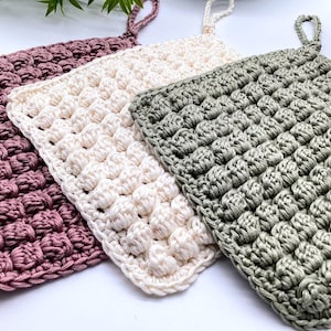 May include: Three crocheted dishcloths in different colors: pink, white, and green. The dishcloths are made with a textured stitch that gives them a puffy appearance.