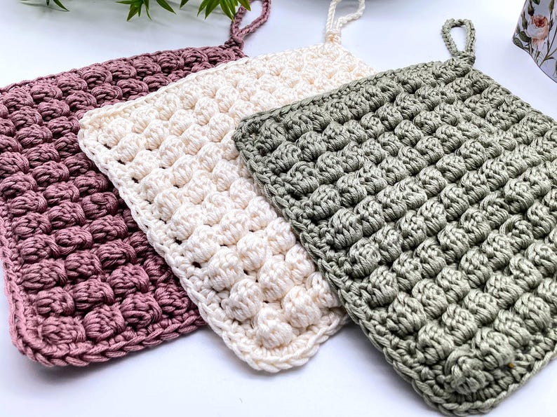 May include: Three handmade crochet potholders in shades of mauve, cream, and sage green. Each square potholder features a textured, raised stitch pattern and a loop for hanging. The potholders are displayed on a white surface.