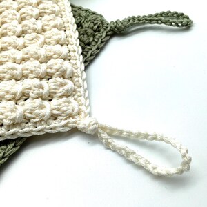 May include: Close-up of two handmade crochet washcloths. One is off-white with a textured, bobble stitch pattern, and the other is olive green. Both have a loop for hanging. The washcloths are made of soft, natural fiber.