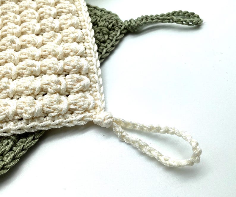 May include: Close-up of two handmade crochet washcloths. One is off-white with a textured, bobble stitch pattern, and the other is olive green. Both have a loop for hanging. The washcloths are made of soft, natural fiber.
