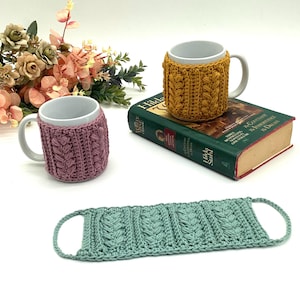 May include: Three white ceramic mugs with decorative, knitted cozies in mustard yellow and mauve. A matching seafoam green coaster is also present. The mugs are on a white surface with a book and floral arrangement in the background.