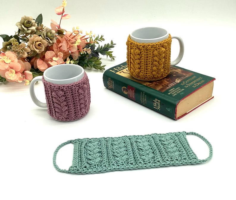 May include: Three white ceramic mugs with decorative, knitted cozies in mustard yellow and mauve. A matching seafoam green coaster is also present. The mugs are on a white surface with a book and floral arrangement in the background.