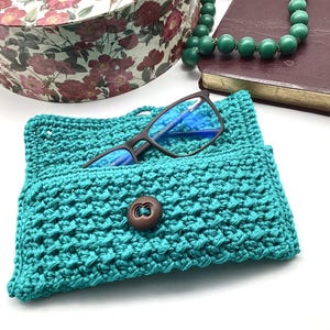 Könnte beinhalten: Türkisfarbene gehäkelte Brillenhülle mit braunem Knopfverschluss. Die Hülle enthält eine Brille mit Blaulichtfilter.