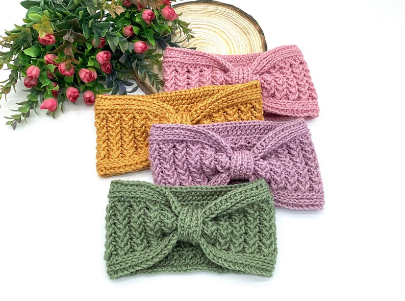 May include: Five knitted headbands in shades of dusty rose, mustard, lavender, and olive green. Each headband has a twisted knot design and a textured knit pattern. The headbands are displayed on a white surface.