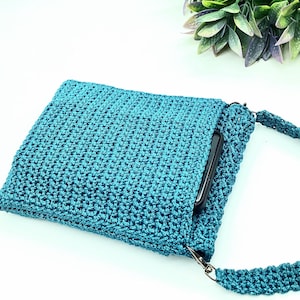 Pu&ograve; includere: Borsa a tracolla all'uncinetto color teal con un motivo a punto strutturato. La borsa ha una forma rettangolare con chiusura a patta e una lunga tracolla abbinata. Uno smartphone nero &egrave; parzialmente visibile all'interno.