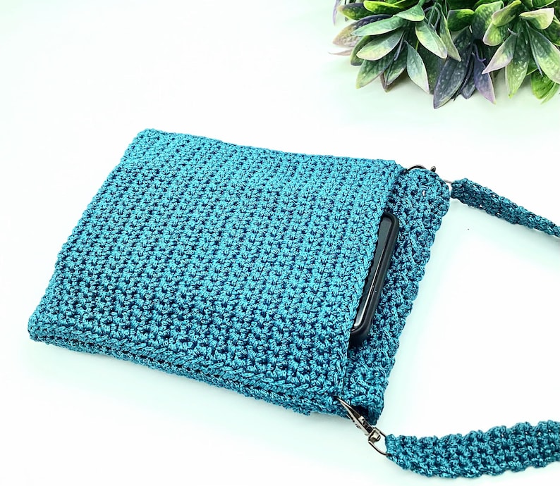 Pu&ograve; includere: Borsa a tracolla all'uncinetto color teal con un motivo a punto strutturato. La borsa ha una forma rettangolare con chiusura a patta e una lunga tracolla abbinata. Uno smartphone nero &egrave; parzialmente visibile all'interno.