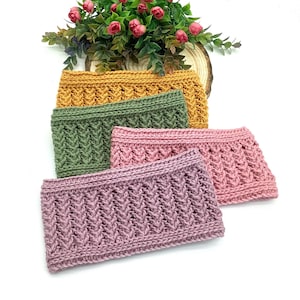 May include: Four knitted headbands in mustard yellow, olive green, light pink, and mauve. Each headband features a textured, woven pattern. A small arrangement of pink flowers and greenery sits in the background.