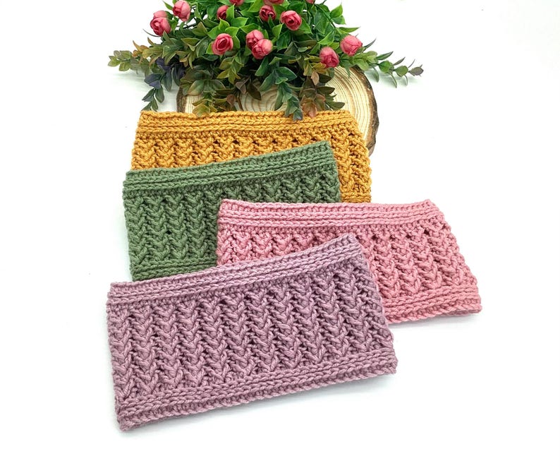 May include: Four knitted headbands in mustard yellow, olive green, light pink, and mauve. Each headband features a textured, woven pattern. A small arrangement of pink flowers and greenery sits in the background.