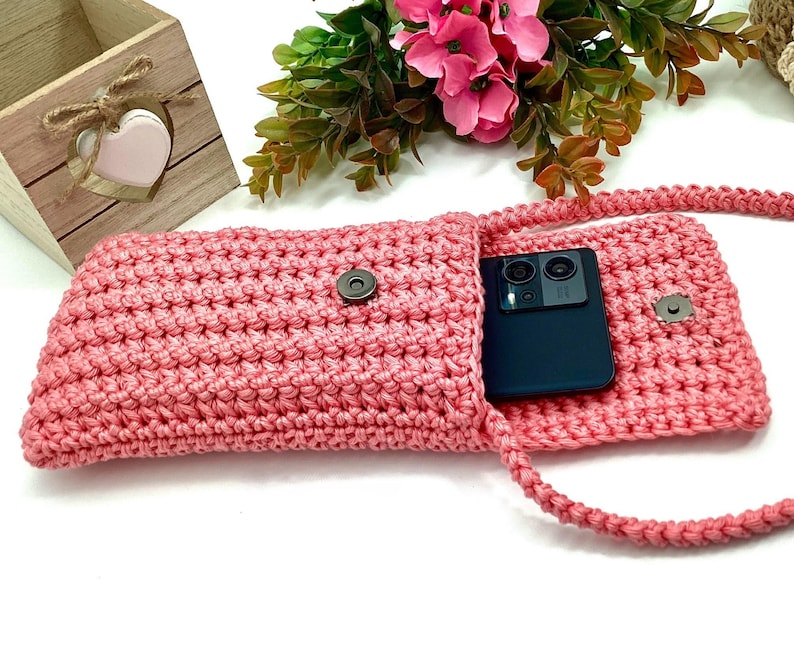 May include: A pink crocheted phone pouch with a snap closure and a long strap. A black smartphone is inside the pouch.