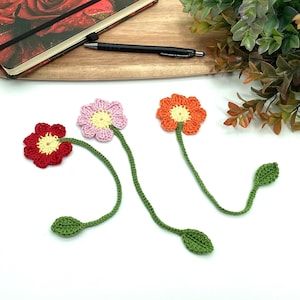 May include: Three handmade crochet flower bookmarks in red, pink, and orange with yellow centers. Each flower has a green stem and leaf. A notebook and pen are in the background.