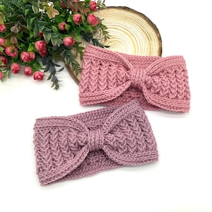 May include: Two knitted headbands in shades of pink and mauve. Each headband features a textured pattern and a bow detail at the centre. The headbands are displayed on a white surface with floral accents in the background.
