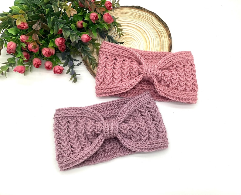 May include: Two knitted headbands in shades of pink and mauve. Each headband features a textured pattern and a bow detail at the centre. The headbands are displayed on a white surface with floral accents in the background.