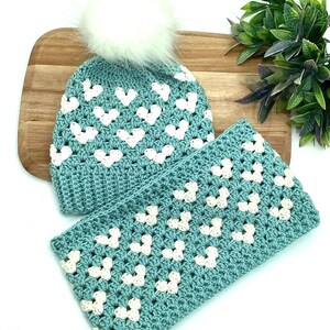 K&ouml;nnte beinhalten: Eine tealblaue H&auml;kelm&uuml;tze mit einem wei&szlig;en Bommel und einem passenden tealblauen H&auml;kelschal mit wei&szlig;en Herzen. M&uuml;tze und Schal sind mit einem Granny-Square-Stich gearbeitet.