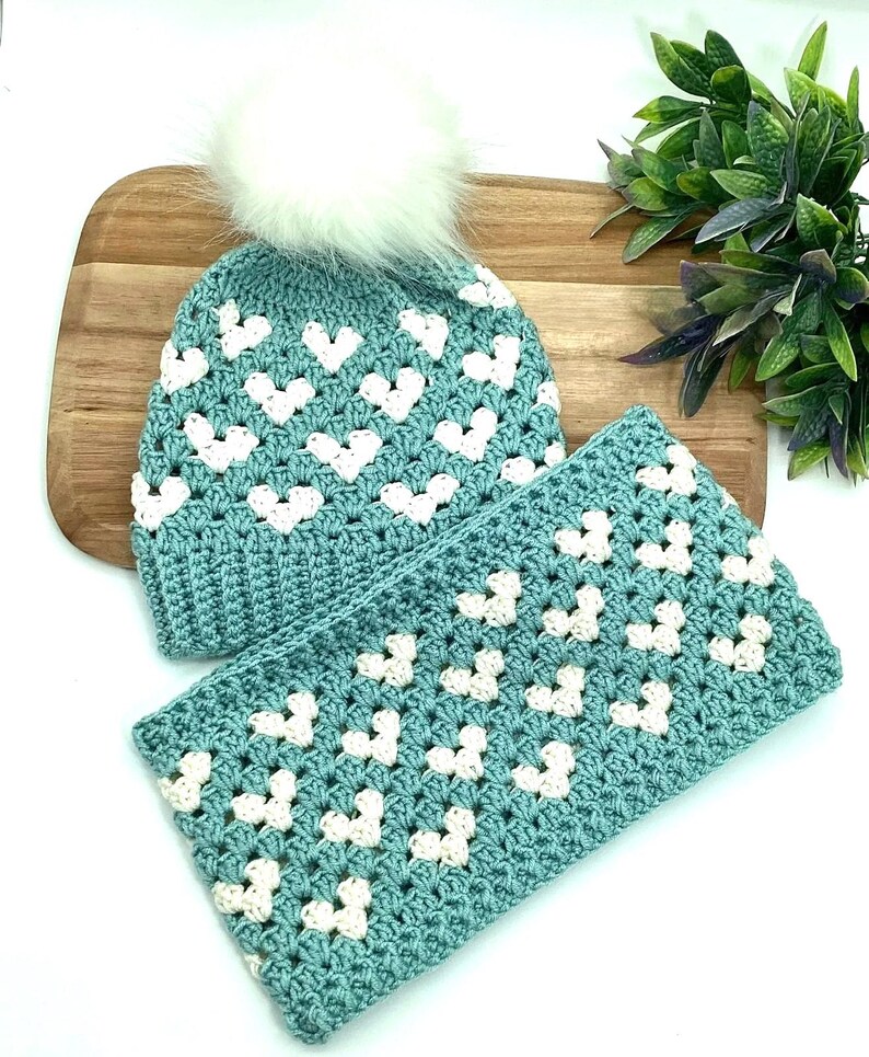 K&ouml;nnte beinhalten: Eine tealblaue H&auml;kelm&uuml;tze mit einem wei&szlig;en Bommel und einem passenden tealblauen H&auml;kelschal mit wei&szlig;en Herzen. M&uuml;tze und Schal sind mit einem Granny-Square-Stich gearbeitet.
