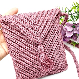 May include: A dusty rose crochet crossbody bag with a textured, diagonal stitch pattern. The bag features a flap closure, a matching tassel, and a braided strap. The handmade bag is a soft pink color.