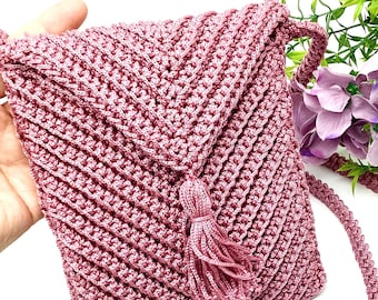 Könnte beinhalten: Eine handgefertigte, altrosa gehäkelte Umhängetasche mit einem strukturierten, diagonalen Stichmuster. Die Tasche hat einen Klappenverschluss, einen passenden Quastenanhänger und einen geflochtenen Riemen. Die Tasche ist rosa.