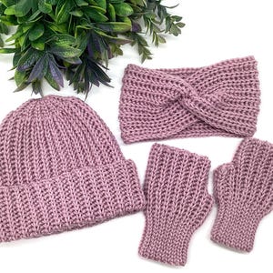 May include: A mauve-colored knitted hat, headband, and fingerless gloves set. The hat has a folded brim. The headband has a twisted design. The fingerless gloves are ribbed. The set is displayed on a white surface with green foliage.
