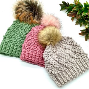 May include: Three handmade crochet beanies in shades of green, pink, and gray, each topped with a fluffy faux fur pom-pom. The beanies have a textured stitch pattern and are arranged in a row, showcasing their colors and textures.