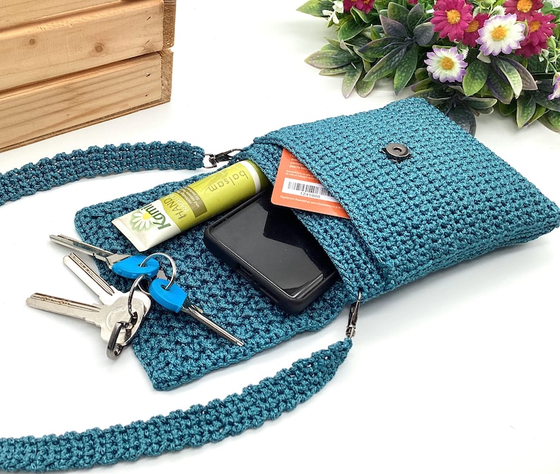 Pu&ograve; includere: Borsa a tracolla all'uncinetto color teal con chiusura a patta, contenente uno smartphone, chiavi e crema per le mani. La borsa ha una tracolla abbinata ed &egrave; aperta, rivelando il contenuto. Sfondo bianco con cassa di legno e fiori.