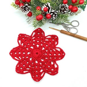 K&ouml;nnte beinhalten: Ein handgefertigter roter H&auml;kelschneeflocke. Die Schneeflocke befindet sich im Vordergrund, mit einer weihnachtlichen Anordnung aus Tannennadeln, roten Beeren und Tannenzapfen im Hintergrund. Ein H&auml;kelhaken und eine kleine Schere sind ebenfalls sichtbar.