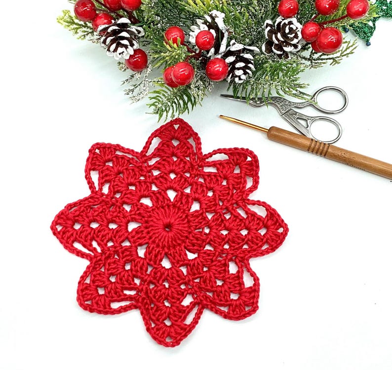 K&ouml;nnte beinhalten: Ein handgefertigter roter H&auml;kelschneeflocke. Die Schneeflocke befindet sich im Vordergrund, mit einer weihnachtlichen Anordnung aus Tannennadeln, roten Beeren und Tannenzapfen im Hintergrund. Ein H&auml;kelhaken und eine kleine Schere sind ebenfalls sichtbar.