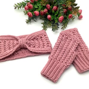 May include: A pink crocheted headband with a bow detail and a pair of matching fingerless gloves. The set is made of a textured yarn and is displayed on a white surface with a floral arrangement in the background.