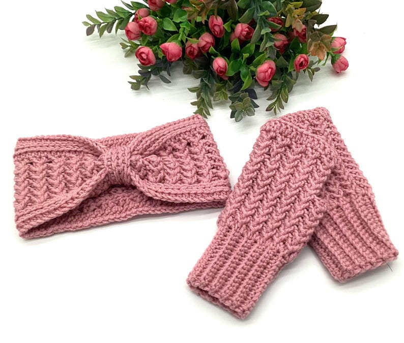 May include: A pink crocheted headband with a bow detail and a pair of matching fingerless gloves. The set is made of a textured yarn and is displayed on a white surface with a floral arrangement in the background.