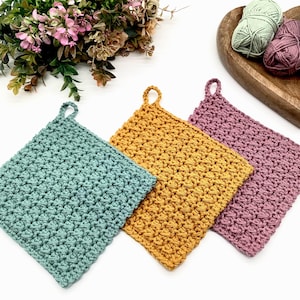 以下が含まれることがあります： ターコイズ、マスタードイエロー、モーヴの色合いの手作りのかぎ針編みの鍋つかみ3つ。各正方形の鍋つかみは、テクスチャードステッチパターンと吊り下げ用のループが特徴です。木製のハート型のボウルに入った毛糸玉が見えます。