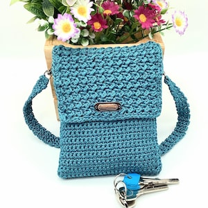 Pu&ograve; includere: Borsa a tracolla all'uncinetto turchese con patta e tracolla. La borsa presenta un punto strutturato e una piccola targhetta metallica con la scritta "Handmade". Un mazzo di chiavi con copri chiavi blu &egrave; davanti.