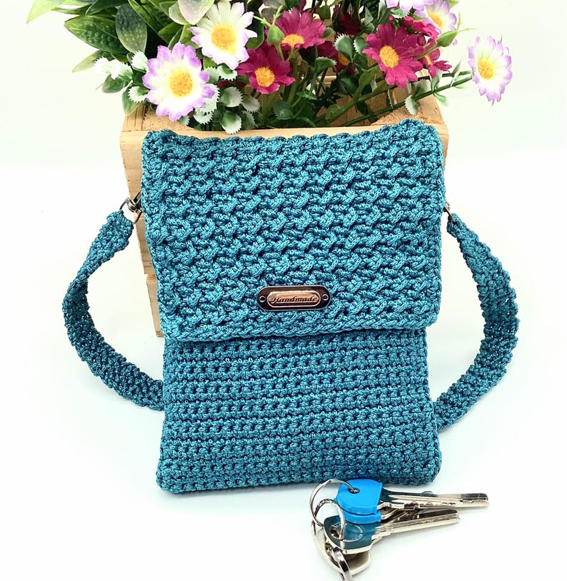 Pu&ograve; includere: Borsa a tracolla all'uncinetto turchese con patta e tracolla. La borsa presenta un punto strutturato e una piccola targhetta metallica con la scritta "Handmade". Un mazzo di chiavi con copri chiavi blu &egrave; davanti.