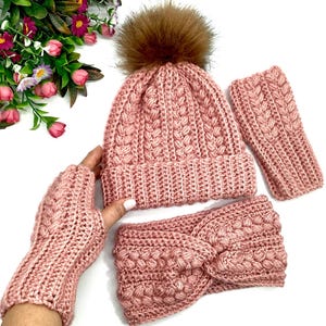 May include: A pink crocheted hat, headband and fingerless gloves set. The hat has a brown faux fur pom pom.