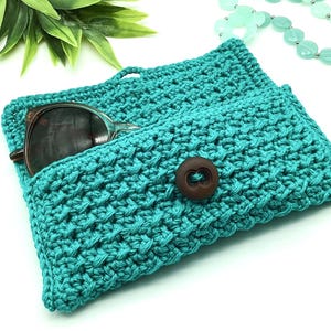 May include: Teal crocheted sunglasses case with a brown button closure. The case is open and a pair of sunglasses is inside.