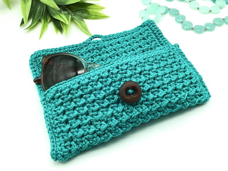 May include: Teal crocheted sunglasses case with a brown button closure. The case is open and a pair of sunglasses is inside.