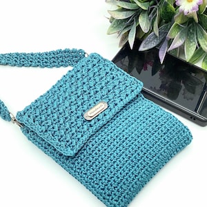 Pu&ograve; includere: Borsa a tracolla all'uncinetto turchese con chiusura a patta e cinturino abbinato. La borsa presenta un motivo a punto strutturato e una piccola targhetta metallica con la scritta "Handmade". Uno smartphone nero &egrave; nelle vicinanze.