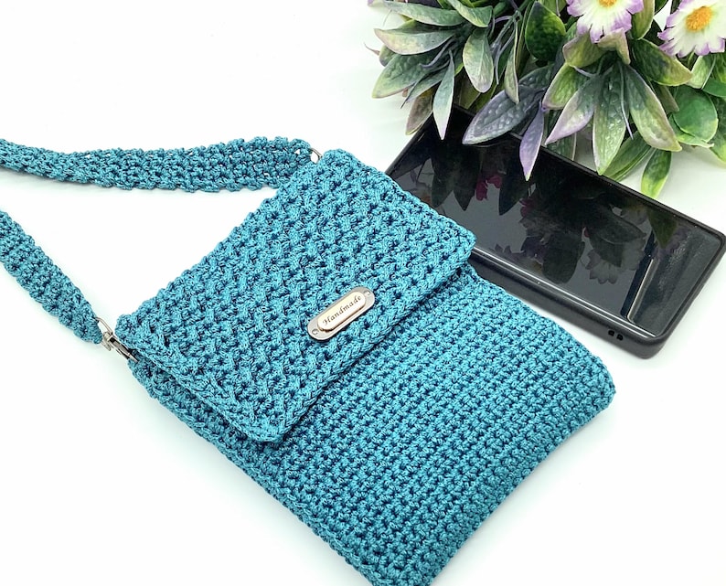 Pu&ograve; includere: Borsa a tracolla all'uncinetto turchese con chiusura a patta e cinturino abbinato. La borsa presenta un motivo a punto strutturato e una piccola targhetta metallica con la scritta "Handmade". Uno smartphone nero &egrave; nelle vicinanze.