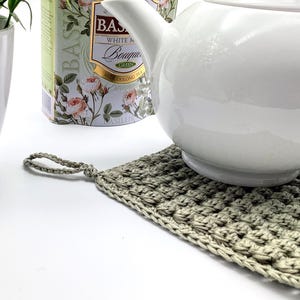 May include: A white ceramic teapot rests on a handmade, square, crocheted trivet. The trivet is a muted green color and has a loop for hanging. A white ceramic pot with a green plant and a tea tin are in the background.