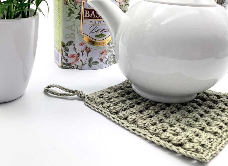 May include: A white ceramic teapot rests on a handmade, square, crocheted trivet. The trivet is a muted green color and has a loop for hanging. A white ceramic pot with a green plant and a tea tin are in the background.